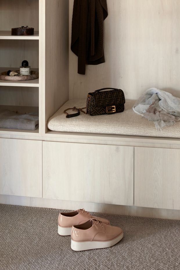 Close-up view of a built-in light timber wardrobe or dressing-room storage unit. Open shelving displays small personal items, folded textiles and accessories, while a hanging rail holds dark clothing. A cushioned beige bench is integrated into the joinery, with shoes placed on the carpeted floor in front.