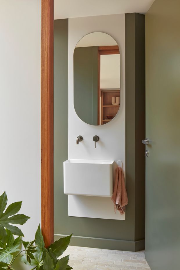 A compact, refined bathroom vanity area viewed through a timber-framed doorway. The space features a wall-mounted white basin with exposed brushed-metal tapware, an arched vertical mirror, deep muted green wall panels, warm timber door and mirror framing, and pale textured flooring. A partially open green door and a small towel add context, while leafy planting is visible in the foreground.
