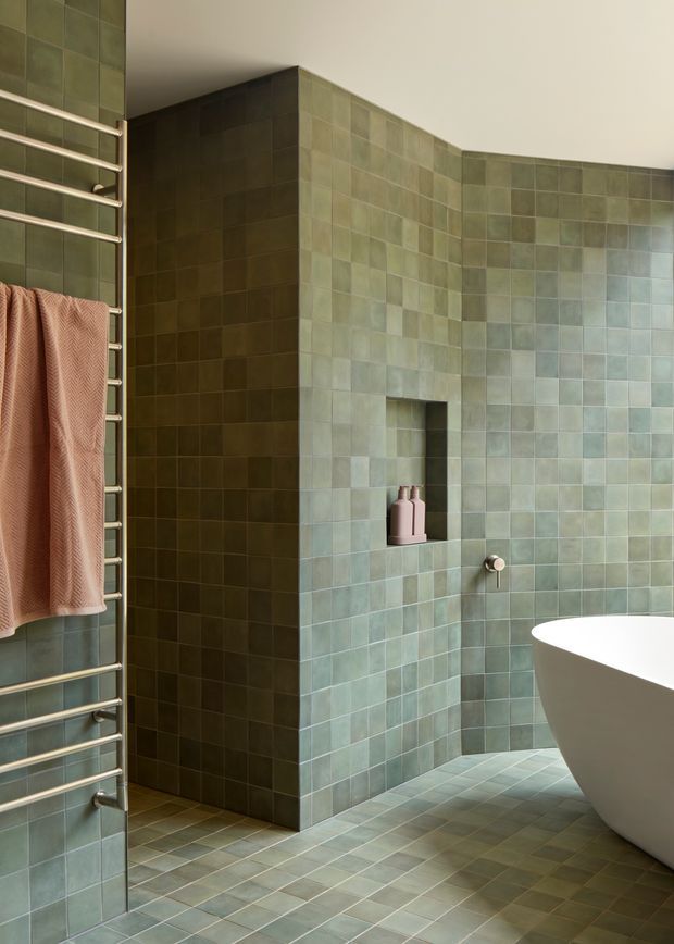 A contemporary bathroom clad in small square green-grey tiles across the walls and floor. A freestanding white bathtub occupies the right foreground, while a recessed wall niche, wall-mounted tap, and stainless-steel heated towel rail with a peach towel are visible. The space has a calm, monochromatic material palette and clean built-in geometry.