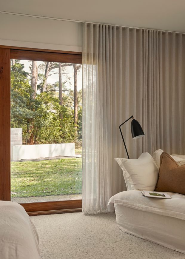 A softly lit bedroom with a large timber-framed glazed opening to a leafy garden. The room features floor-to-ceiling sheer curtains, a plush white upholstered bed or daybed with neutral cushions, a black angled floor lamp, and pale carpet. The composition emphasizes the connection between the calm interior and the wooded outdoor setting.