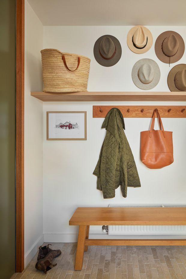 A compact, warmly styled entryway with a built-in timber coat rack and upper display shelf. Woven baskets, wall-mounted hats, a framed print, a quilted jacket, leather tote, and timber bench create an organised mudroom-like entrance. The space has pale walls, warm timber trim and joinery, and a narrow light stone- or concrete-look tiled floor.