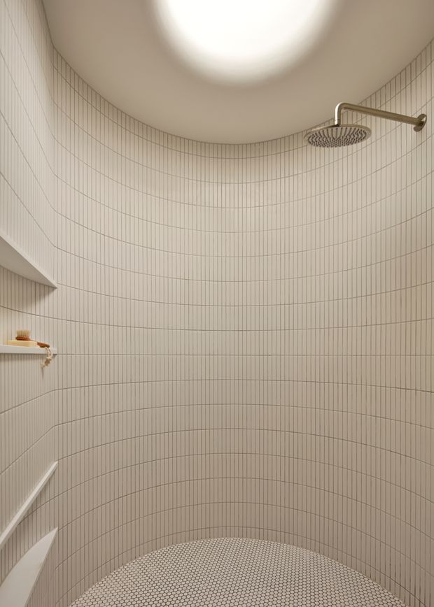 Curved walk-in shower enclosure with fully tiled walls in a narrow vertical grid pattern and a small hexagonal mosaic tile floor. A wall-mounted rainfall shower and recessed corner shelf are visible. The rounded ceiling includes a large circular skylight or ceiling light, creating a soft, warm and minimal atmosphere.