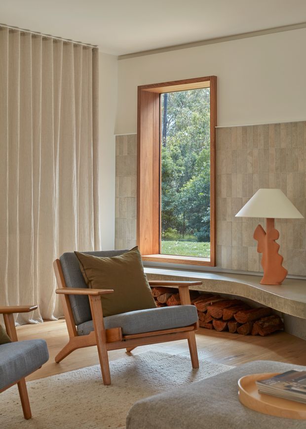 A warm, refined living room with a mid-century-inspired timber lounge chair, neutral upholstered seating, full-height beige curtains, and a built-in tiled fireplace hearth with stacked firewood beneath. A tall timber-framed window opens the room to leafy greenery outside. A sculptural orange table lamp and soft neutral furnishings add understated accents.