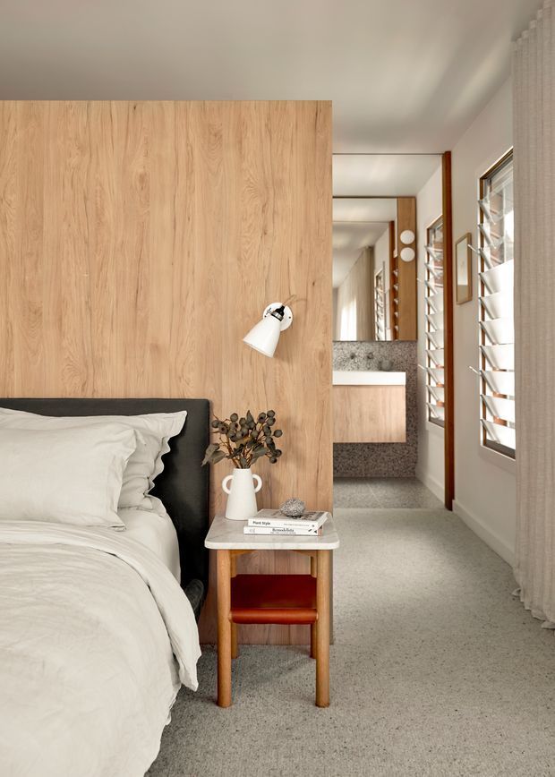 A warm, refined bedroom with a partial-height light timber feature wall behind a bed, integrated white wall light, and a compact timber bedside table. The room extends toward a bathroom or vanity area with a stone-textured wall and basin visible in the background. White bedding, pale walls, grey carpet, timber trims, framed artwork, and louvred windows create a calm contemporary interior.