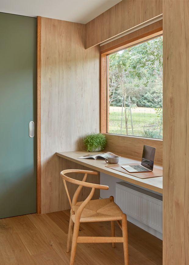 A compact built-in home workspace set within a warm timber-lined alcove. The space features a wall-to-wall timber desk beneath a framed window, a laptop, books, plant and cup, with a sculptural wooden chair in front. A muted green sliding door and radiator add contrasting elements. The interior is calm, functional and strongly defined by natural timber joinery and flooring.