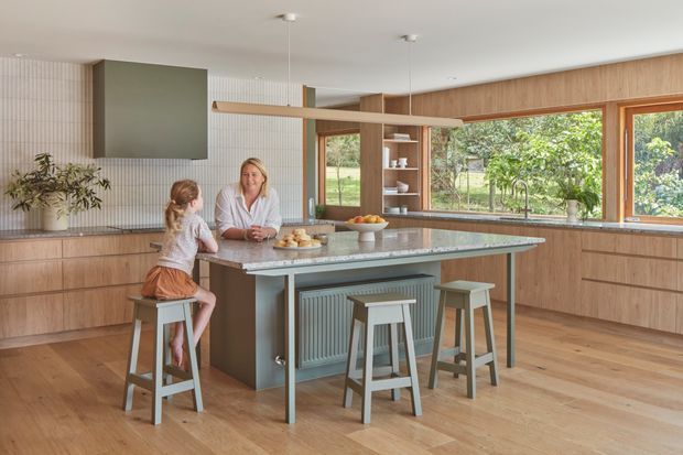 A bright, spacious contemporary kitchen with extensive light timber cabinetry, a large stone island with integrated seating, and full-height corner glazing overlooking greenery. A tiled feature wall, suspended timber shelf, open shelving, and muted sage-green accents create a warm, family-oriented space.