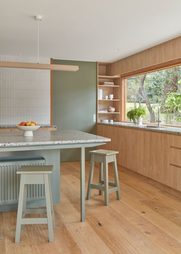 A warm, contemporary kitchen with extensive light timber cabinetry, a pale stone island benchtop, muted sage-green wall panels, and integrated open shelving. Two stools sit at the island, while a large timber-framed window above the sink looks onto leafy gardens. The room features timber flooring, a slim linear pendant, ribbed wall detailing, and a calm natural palette.