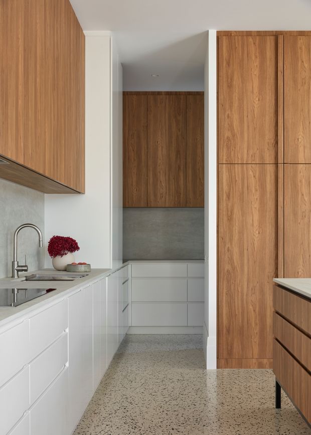 A narrow contemporary kitchen with a galley-style layout. The room features white lower cabinetry, extensive warm timber cabinetry and wall panels, a grey concrete-look backsplash, a stainless steel sink and faucet, and a speckled terrazzo or concrete-look floor. A peninsula or island with a matching speckled benchtop occupies the foreground. The space is clean, restrained, and materially focused.