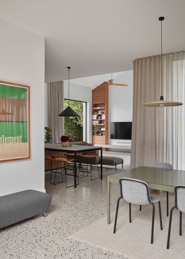 A bright, contemporary open-plan living and dining interior. The foreground has a muted green dining table with upholstered grey chairs on a light rug, while the adjoining space contains a dark-framed dining table, timber seating, a grey sofa, built-in timber shelving and a wall-mounted television. Full-height sheer curtains, pendant lights, a timber ceiling fan and large windows contribute to the airy, refined atmosphere. The polished terrazzo floor extends throughout the room, with white walls and a framed artwork adding visual interest.