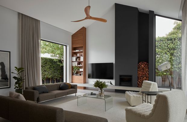 A refined contemporary living room with a high sloping ceiling, extensive glazing, neutral furnishings, and a dramatic black fireplace wall. The room includes a sectional sofa, sculptural lounge chairs, a glass coffee table, built-in timber shelving, a television, indoor planting, and a visible outdoor terrace beyond the glazing. A timber ceiling fan adds a warm natural accent.