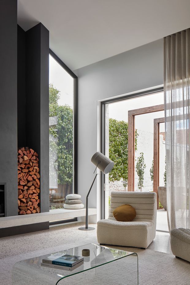 A refined contemporary living room with a dramatic black fireplace and integrated firewood storage, expansive corner glazing, and a glazed opening to a landscaped courtyard. The room features pale textured upholstery, a sculptural floor lamp, sheer curtains, a glass coffee table, and a soft neutral palette. Timber-framed courtyard elements and greenery are visible outside.