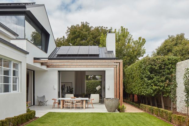 Exterior view of a contemporary residential extension opening onto a landscaped courtyard lawn. The white rendered house has large dark-framed windows, a flat-to-low-pitched roof with visible solar panels, and a timber-framed covered outdoor dining area. A glazed opening reveals an interior dining or living space, while established hedging and trees create a private garden setting.