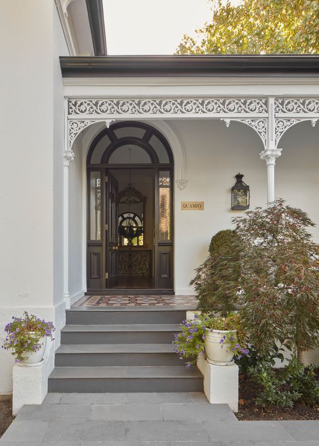 A formal heritage-style building entrance viewed from outside, with broad grey stone steps leading to a dark timber arched doorway. The entry porch features ornate white cast-iron lacework, slender columns with decorative capitals, intricate trim, a patterned tiled threshold, and white rendered walls. Potted flowering plants and established shrubs frame the entrance, while a small sign reads “QUAMBY.”