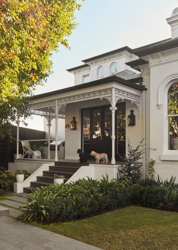 A sunlit front exterior of an ornate heritage-style house, featuring a covered verandah with decorative cast-iron lacework, slender columns, dark timber steps, double entry doors, arched windows, classical cornices, and a carefully landscaped garden. A small dog and outdoor furniture are present, with a sidewalk and lawn in the foreground.