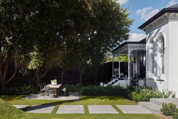 A landscaped residential courtyard beside a white heritage-style house, featuring ornate arched wall detailing, decorative porch fretwork, dark-framed glazing, clipped planting beds, a paved outdoor dining area, and large rectangular stepping stones set into the lawn. Mature trees provide shade and contrast against the bright facade.