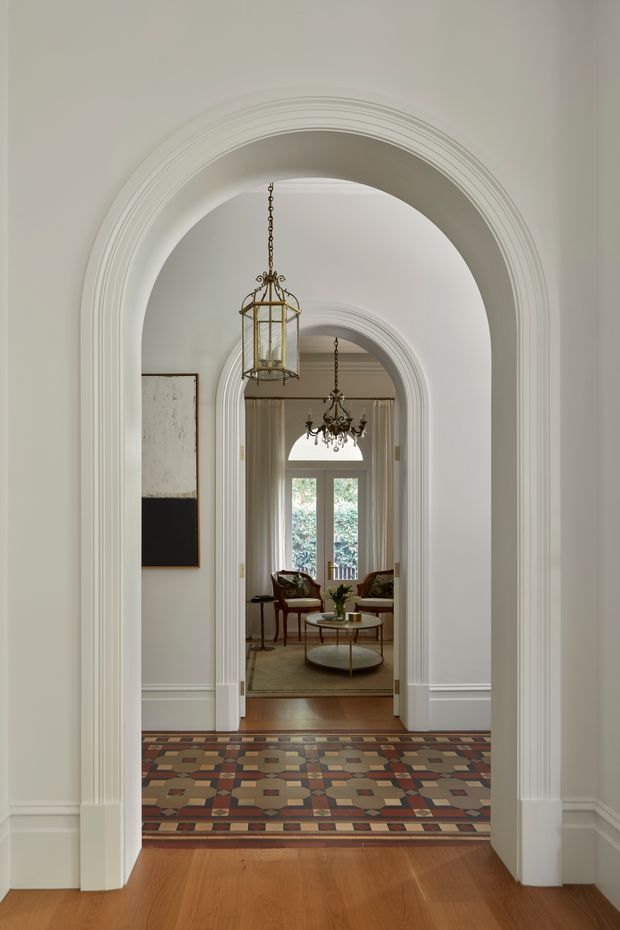 A refined interior hallway viewed through a series of decorative white arched openings with layered moulded trim. The foreground has warm timber flooring, while a colourful geometric patterned tiled or mosaic floor leads toward a sitting room. Ornate hanging lanterns and a chandelier emphasize the traditional character, with upholstered armchairs, a round coffee table, tall glazed doors, and restrained artwork visible in the background.