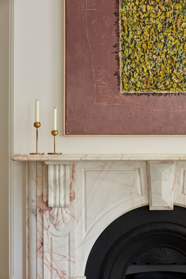 Close-up interior detail of an ornate fireplace mantel, carved pale marble with prominent red veining and classical fluted detailing. Two slender brass-and-cream candlesticks sit on the mantel, while layered abstract artwork in muted burgundy and textured green-yellow tones hangs above. The composition has a refined, historic character with warm neutral wall colouring.