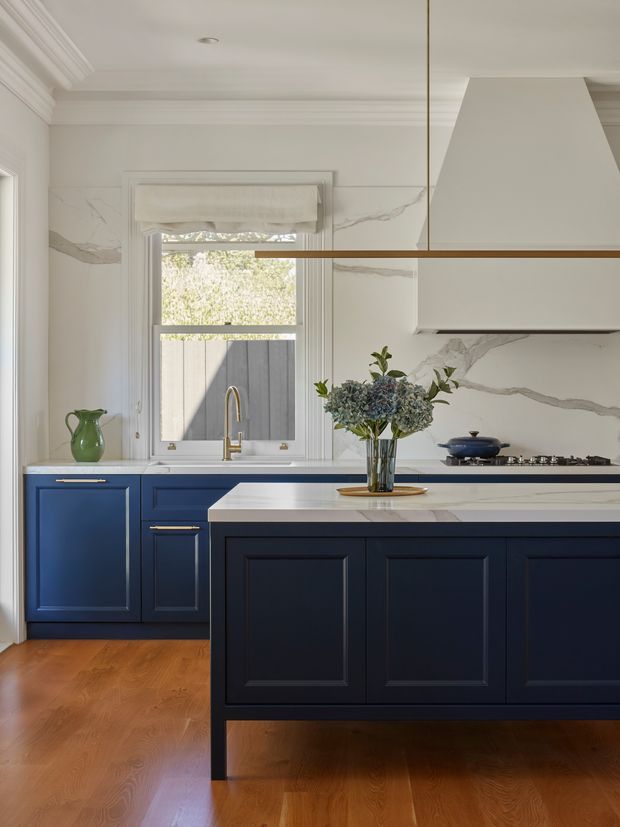 A refined traditional kitchen with deep navy-blue shaker cabinetry, marble-look veined wall surfaces, and a pale stone island benchtop. The room features ornate ceiling cornices, framed window trim, brass hardware and tapware, a linear brass pendant, timber flooring, and a large white rangehood. A vase of foliage and blue cookware add restrained decorative detail.