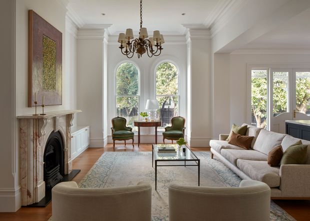 Elegant, light-filled formal living room with high ceilings, ornate white wall mouldings, a decorative marble fireplace, tall arched windows, and classic furnishings. A large sectional sofa, curved armchairs, glass coffee table, chandelier, and muted patterned rug create a refined traditional interior.