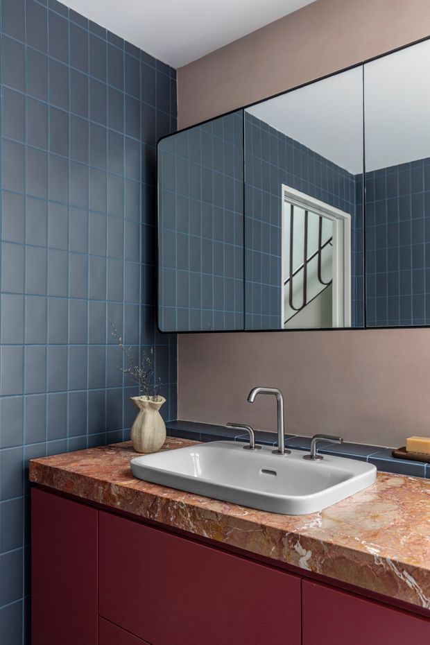 A stylish bathroom vanity featuring a white vessel sink with chrome tapware, a richly veined reddish-brown stone benchtop, and deep burgundy fixed cabinetry. The walls are clad in dark blue square tiles with contrasting grout, while a warm taupe painted wall surrounds a large black-framed mirror. A small sculptural vase and soap add minimal decoration.
