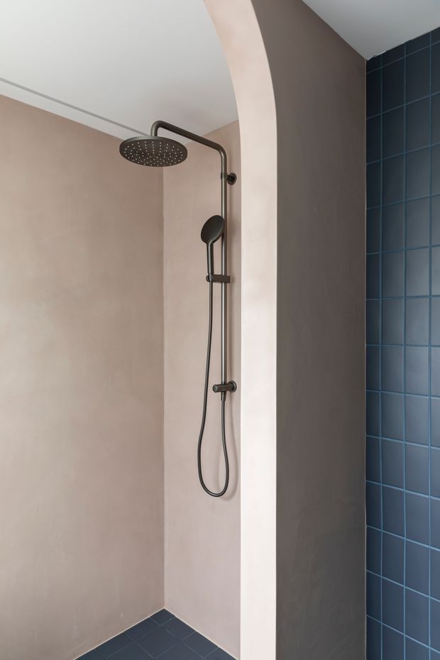 A contemporary walk-in shower with a dark metal rainfall showerhead and handheld fixture mounted against warm taupe plaster walls. A curved plaster partition frames the shower, while deep blue square tiles cover the adjacent wall and floor, creating a strong colour contrast. The space is minimal and uncluttered with clean-lined architectural detailing.