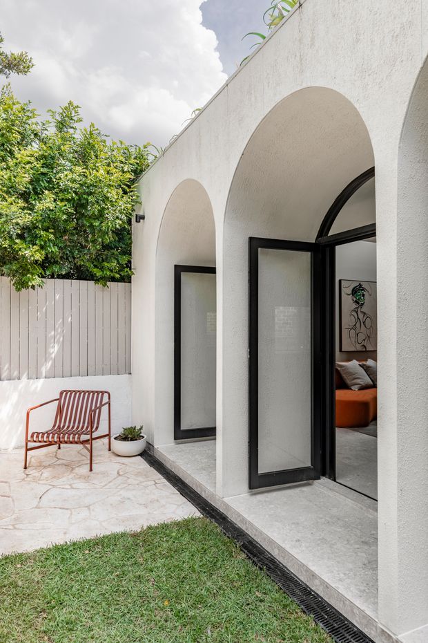 A bright courtyard or patio beside a white textured stucco building with a series of large rounded arches and black-framed glazed doors. One door is open, revealing part of an orange-toned interior seating area. The paved terrace has irregular pale stone flooring, a red striped metal lounge chair, a small potted plant, and a narrow linear drain along the building edge. Lush greenery and a pale vertical fence enclose the space, creating a light Mediterranean-inspired outdoor setting.