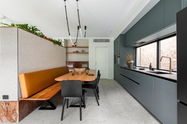 Contemporary open-plan kitchen and dining area with a built-in banquette beside a compact dining table. The room features dark blue-grey full-height cabinetry, a long stone-look benchtop, black-framed windows, polished concrete flooring, warm timber and orange upholstery, and a textured partition wall topped with indoor plants. Linear pendant lighting and integrated joinery create a refined, functional interior.