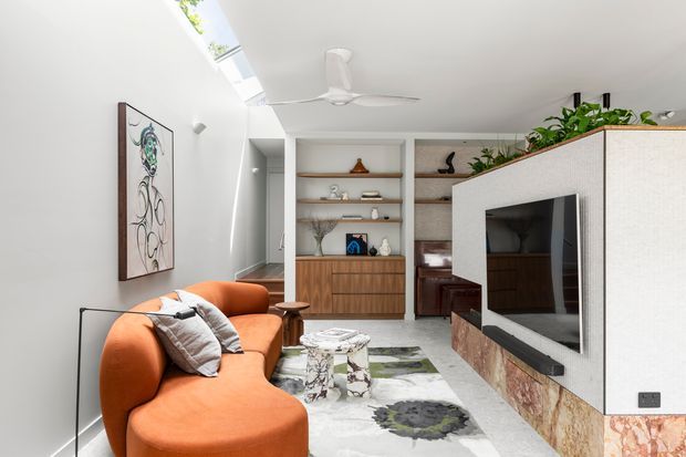 A bright contemporary living room with a curved orange sofa, wall-mounted television set into a textured partition, built-in timber shelving and cabinetry, a patterned area rug, indoor plants, and a skylight with a ceiling fan. A hallway or adjoining circulation space is visible beyond the room.