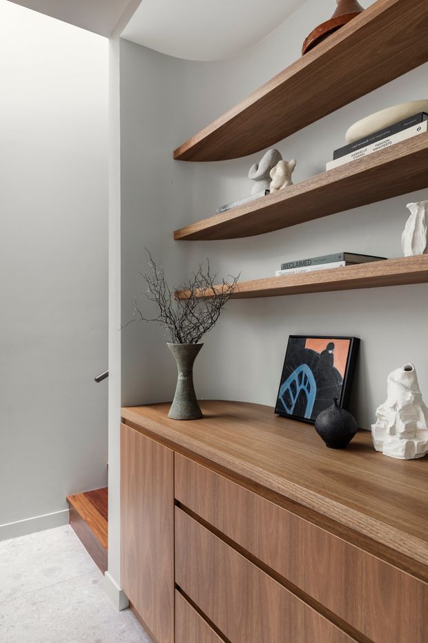 A refined built-in hallway display and storage unit with warm walnut-toned cabinetry, a continuous timber countertop, and three floating timber shelves styled with books, sculptural ceramics, a framed artwork, and a vase of branches. The cabinetry sits beside a stairway, with softly curved white plaster walls and a pale stone or concrete-look floor. The composition is minimalist, warm, and carefully curated.