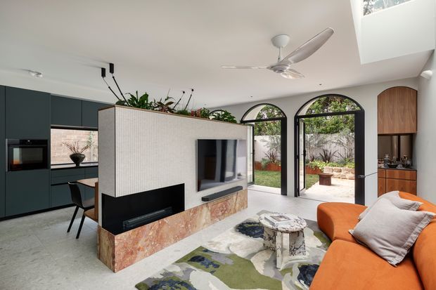 A bright contemporary open-plan living room with a warm orange sectional sofa, sculptural patterned rug, and a central feature wall containing a television and fireplace. Dark built-in kitchen cabinetry is visible to the left, while arched glazed doors open to a landscaped courtyard. The feature wall combines ribbed light textured cladding with a pink-toned stone base, and a timber-toned built-in joinery element frames the right side. A ceiling fan and rooflight add to the airy, light-filled space.