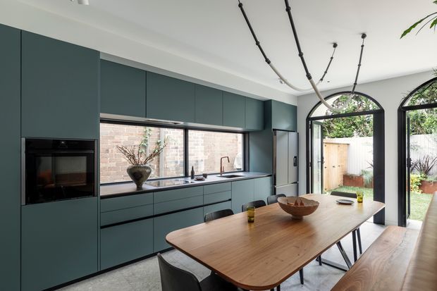 A contemporary open-plan kitchen and dining area with dark teal full-height cabinetry, integrated appliances, a long timber island or dining bench, and a large timber dining table with bench seating. Black-framed arched glazed doors open to a small courtyard, while a sculptural linear pendant hangs from the ceiling. Pale concrete-look flooring and exposed brick visible through the glazing add texture.