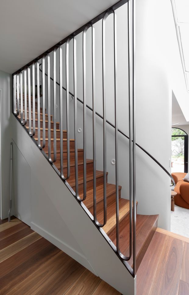A bright contemporary stairway with warm timber treads and flooring, white walls, and a distinctive dark metal balustrade composed of slender vertical rods with rounded U-shaped ends. The stair rises alongside a large light-filled opening, with a glimpse of an adjacent living area at right.