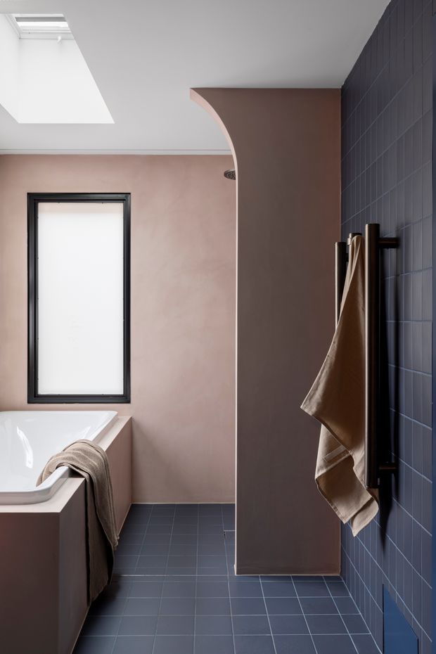 A refined contemporary bathroom with a freestanding white bathtub, soft pink plastered walls, dark blue tiled flooring and a matching dark blue tiled feature wall. A black-framed frosted window and large skylight provide natural light, while a curved plaster arch conceals the shower or vanity area. Dark bronze or black towel hardware and neutral towels add contrast.