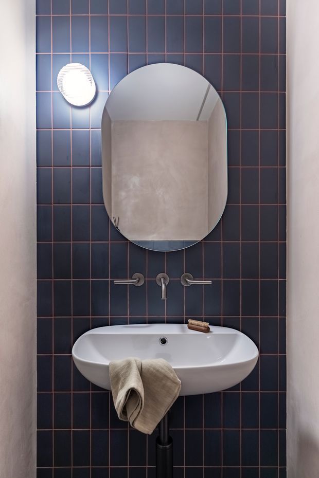A compact bathroom vanity is framed against a dark navy tiled feature wall with contrasting thin copper-toned grout lines. The wall-mounted white ceramic basin has exposed brushed-metal wall-mounted taps, a rounded vertical mirror above, and a small wall light to the upper left. A neutral towel and wooden brush add restrained warmth to the otherwise dark, refined space.
