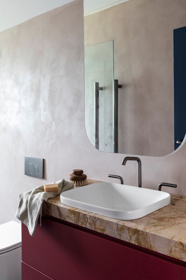 A contemporary bathroom vanity with a white vessel basin, dark brushed-metal wall-mounted tapware, and a large rounded-corner mirror. The vanity features deep burgundy cabinetry and a prominently veined warm marble or natural-stone benchtop. Soft pink-beige textured wall finish, muted towels, and understated accessories create a refined, warm interior.