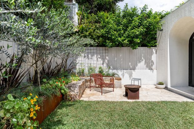 A bright, landscaped courtyard garden with a pale stone-paved sitting area, a rust-coloured woven lounge chair, a small round fire pit, raised planting beds, mature shrubs and a white rendered wall with a large arched opening. The space has a refined Mediterranean-influenced contemporary feel and opens onto a neatly maintained lawn.