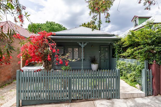 Front exterior view of a compact single-storey heritage-style home with a grey weatherboard facade, corrugated metal hipped roof, small covered verandah, decorative timber posts, and a pale picket fence. Lush established planting and neighbouring homes frame the suburban streetscape.