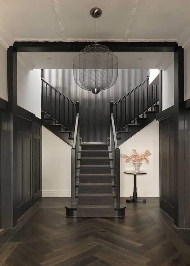 A dramatic, symmetrical interior stair hall viewed from an entry-like corridor. A central carpeted staircase divides into two upper flights with dark railings, set against vertically ribbed dark wall cladding. The space features dark paneled joinery, a dark herringbone timber floor, white walls and ornate ceiling cornices, plus a large wire-frame pendant light and a small round console table with a vase.