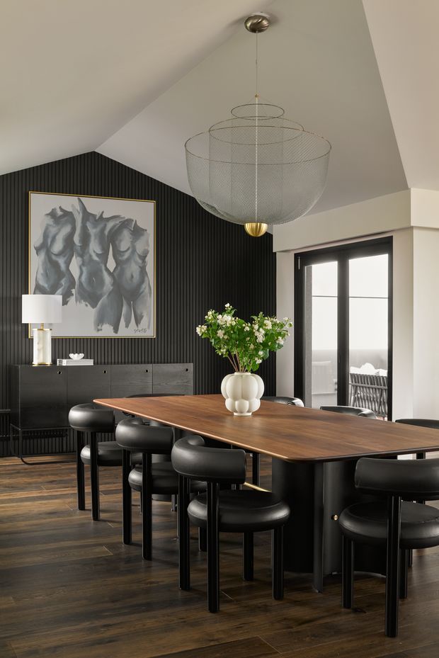 A refined dining room with a long rectangular timber dining table surrounded by black upholstered chairs. The room features dark vertical wall panelling, dark timber flooring, a large abstract artwork, a black sideboard, and a sculptural tiered wire pendant light. Glazed double doors open toward an exterior balcony or terrace. The palette is predominantly dark wood, black, white, and muted grey with warm brass accents.