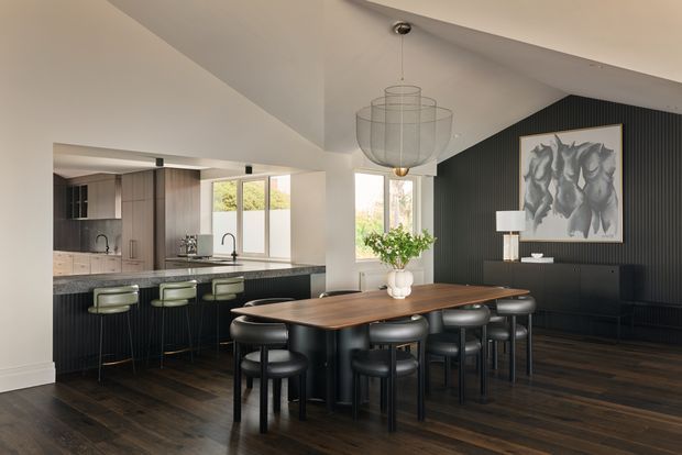 Open-plan dining and kitchen interior with a long dark timber dining table surrounded by black upholstered chairs. The kitchen has dark cabinetry, a stone island benchtop, integrated appliances and stools. A black vertical-panel feature wall, sculptural pendant light, artwork and sideboard create a refined contemporary look, while large windows provide natural light.