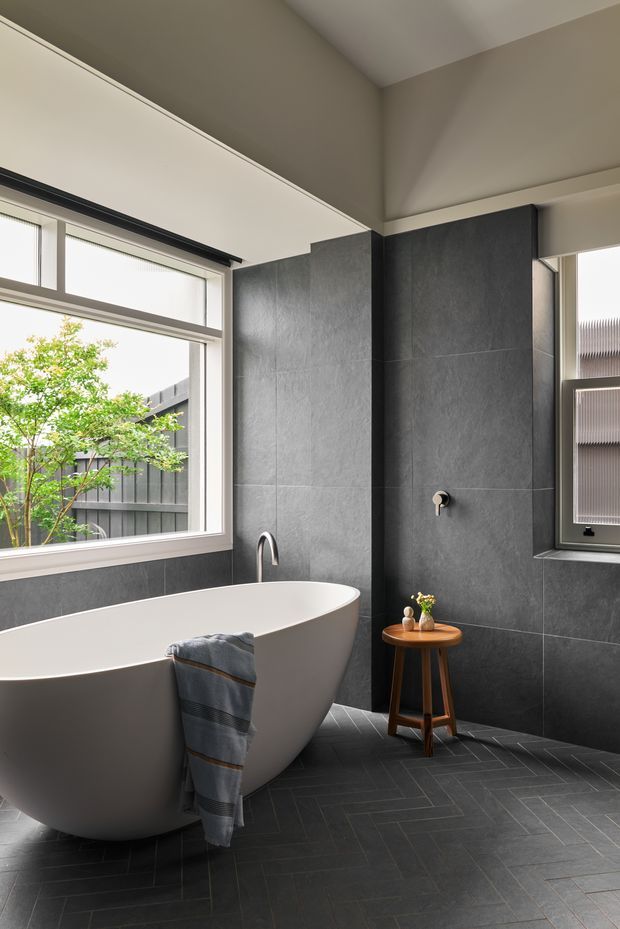 A contemporary bathroom featuring a freestanding white oval bathtub, dark grey tiled walls and floor, a wall-mounted bath fitting, and large windows bringing in natural light. A small timber stool with bathroom accessories sits beside the tub, while a grey towel is draped over its edge.