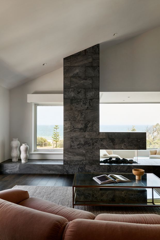 A refined living room with expansive coastal views through large windows. A dramatic dark natural-stone fireplace and hearth form a sculptural floor-to-ceiling feature wall, extending into a horizontal media or display ledge. The room includes a low glass-and-dark-stone coffee table, a large soft textured rug, and a curved blush-toned sofa. Neutral plaster walls, recessed ceiling lights, timber flooring, and minimalist styling create a calm, contemporary interior.