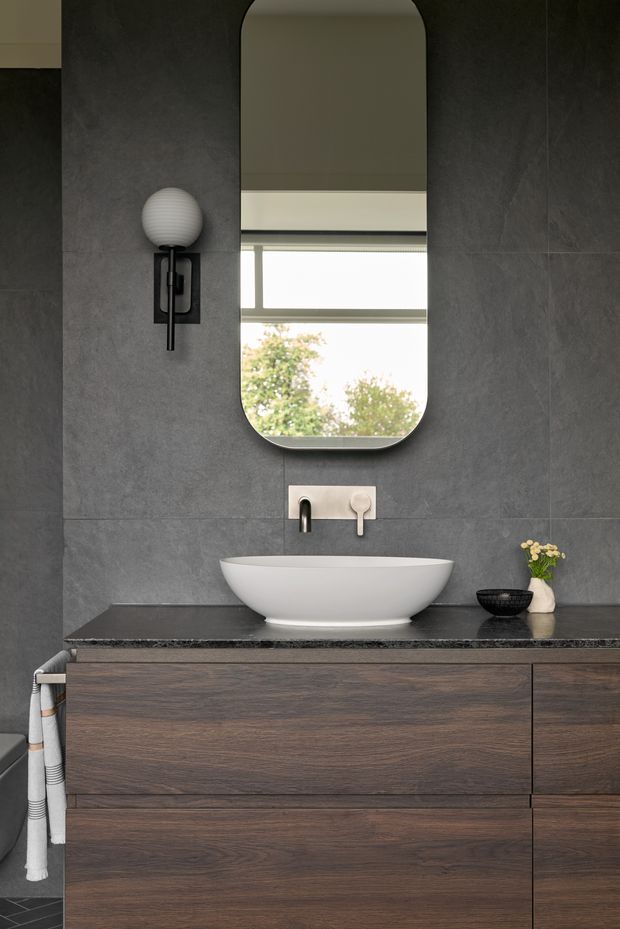 A refined contemporary bathroom vanity featuring dark grey large-format wall tiles, a dark stone benchtop, a white vessel basin, wall-mounted brushed metal tapware, and a warm dark timber floating cabinet. An arched vertical mirror, globe wall sconce, and minimal accessories complete the balanced, moody composition.