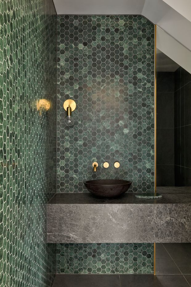 A close-up view of a refined bathroom vanity beneath a sloping ceiling. The walls are clad in small irregular green hexagonal tiles with dark grout, contrasted by a dark stone benchtop and projecting grey stone vanity front. A dark vessel basin, wall-mounted brass tapware, and a round brass wall light create warm accents. A frameless glass shower screen with a slim brass edge is visible to the right, with dark floor and wall tiles continuing into the shower.