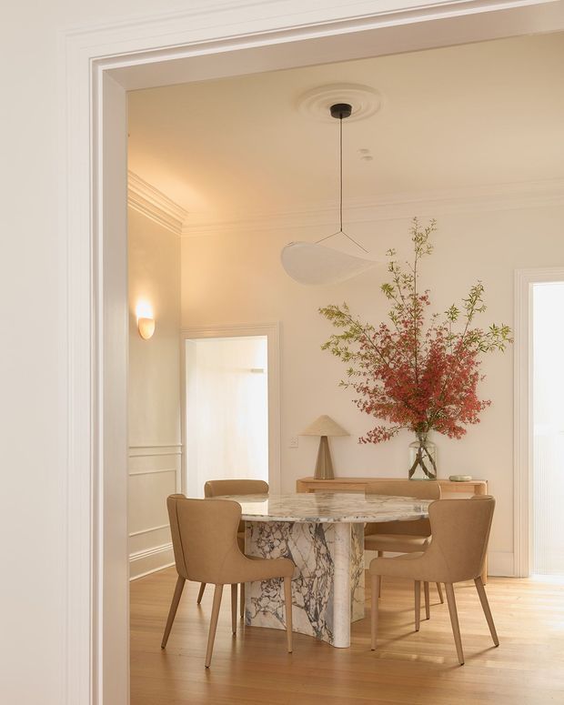 Elegant dining room viewed through a wide, moulded doorway. The room features a round marble dining table with a sculptural veined stone pedestal, four upholstered tan dining chairs, warm timber flooring, cream walls with decorative wall panelling and ornate ceiling cornices, a contemporary pendant light, wall sconces, and a large vase of branches on a timber console. The overall look is refined, warm, and softly traditional with contemporary furnishings.