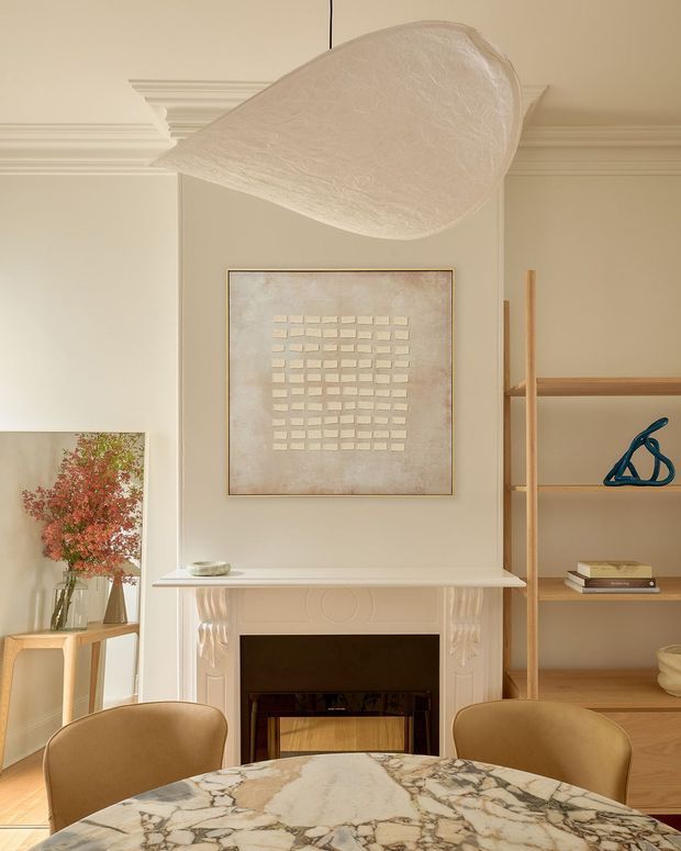 Elegant warm-toned dining and living area centered on a decorative fireplace, with a round veined stone dining table and upholstered dining chairs in the foreground. Built-in open timber shelving, framed artwork, a tall sculptural pendant light, ornate ceiling cornice, and soft neutral finishes create a refined contemporary-classic interior.