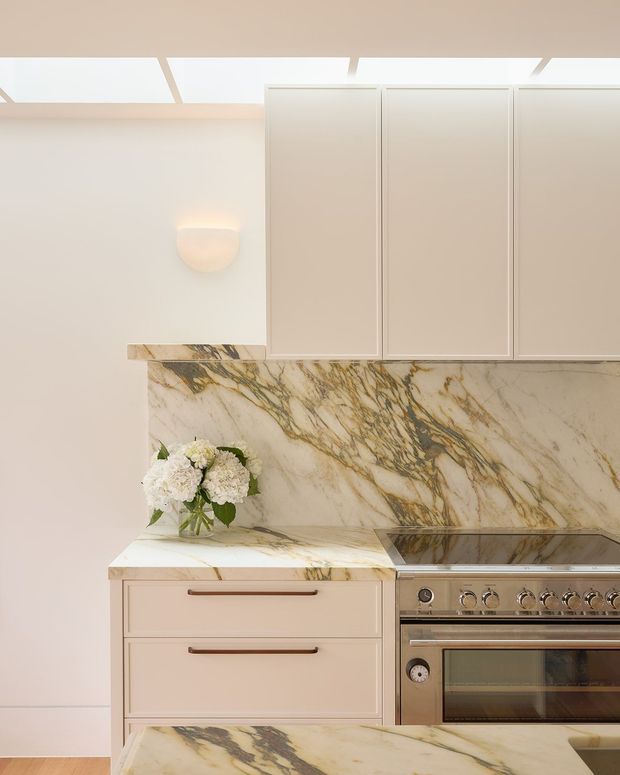 A refined kitchen detail with pale warm cabinetry, long metal drawer pulls, a dramatic veined marble splashback and benchtop, and a stainless-steel range cooker. A skylight-style ceiling opening and soft wall sconce provide diffuse illumination, while a small vase of white flowers adds a decorative touch.