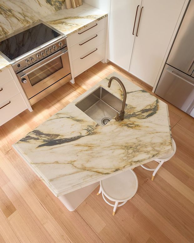High-angle view of a refined kitchen centred on a large marble island with an integrated stainless-steel sink and curved bronze-toned tap. The island has rounded corners and seating beneath it. The kitchen includes a stainless-steel range cooker, white cabinetry with long dark metal handles, a matching marble benchtop and backsplash, and warm timber flooring.
