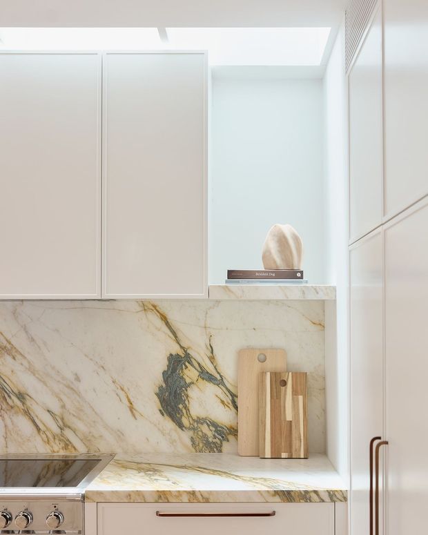 Close-up view of a refined kitchen with pale matte cabinetry, dramatic veined natural stone backsplash and benchtop, integrated cooktop and oven controls, and warm timber cutting boards displayed on the counter. A bright skylight or clerestory opening illuminates the predominantly white space, while slim bronze-toned cabinet handles add contrast.