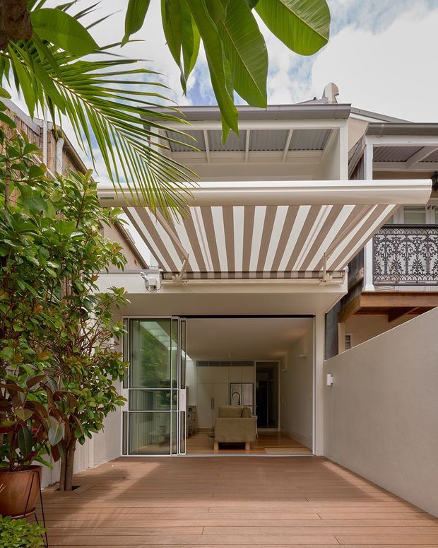 A compact residential courtyard or rear deck viewed toward an open, glazed ground-floor living area. The space features warm timber decking, pale rendered walls, a retractable striped awning, lush tropical planting, and a light-coloured multi-storey facade with ornate metal balcony detailing.